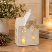 White ceramic tealight house with sculpted beige bow and illuminated windows beside winter greenery