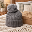 Charcoal cable knit bobble hat styled in a warm bedroom beside a gift box