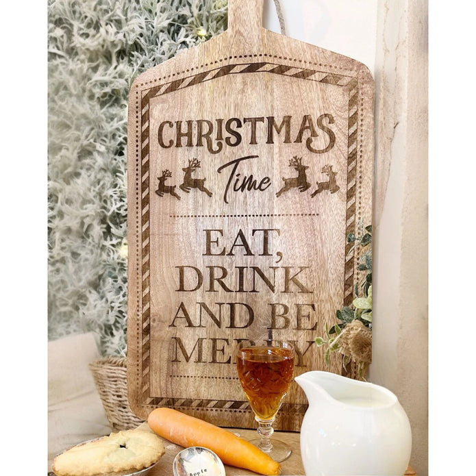 Wooden Christmas serving board with brown Eat, Drink and Be Merry lettering, propped beside cheese and a drink.