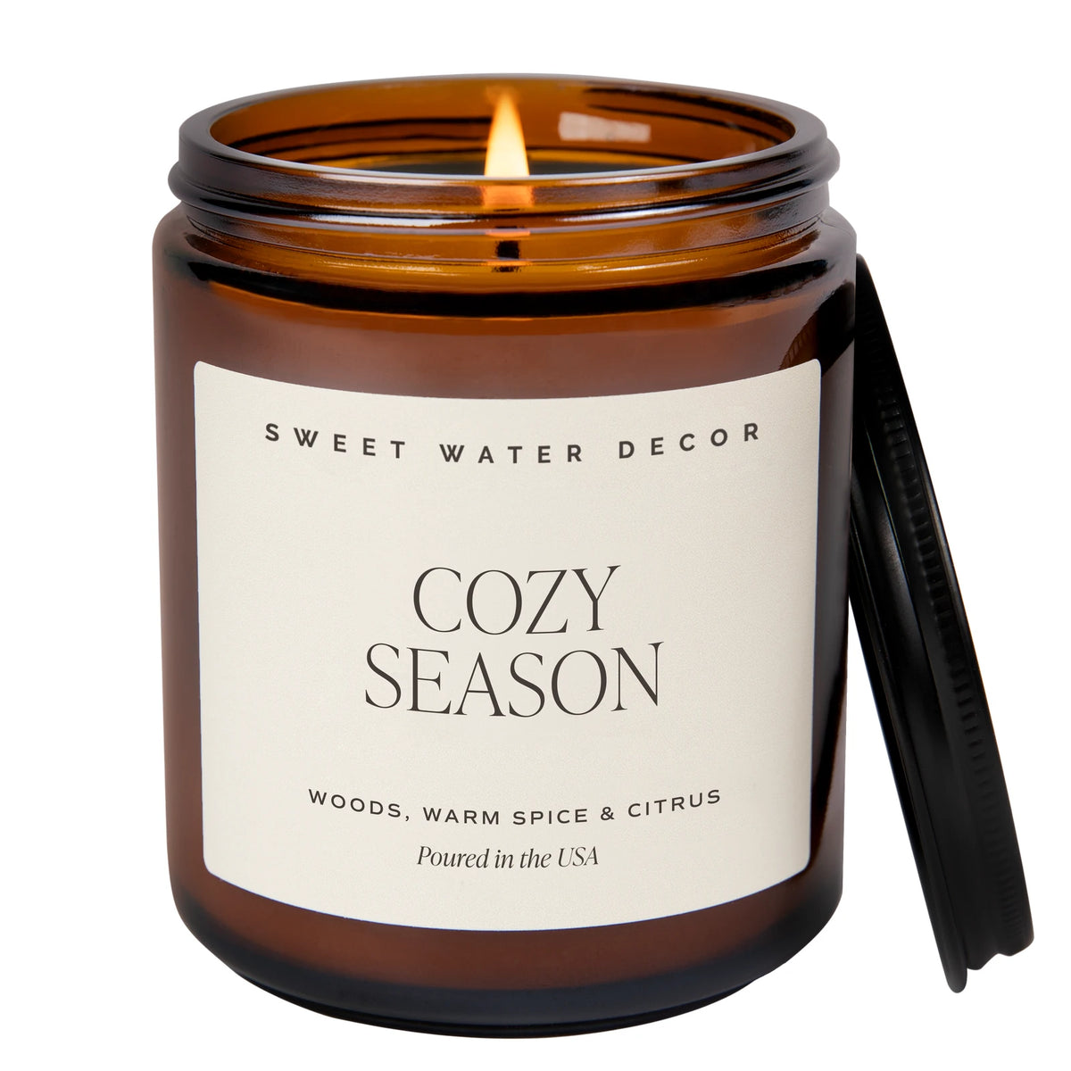 Lit Sweet Water Decor Cozy Season amber candle with its black lid beside the jar on a white background.