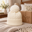 Cream cable knit bobble hat styled in a warm bedroom beside a gift box