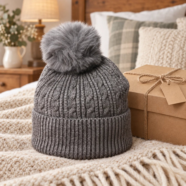 Charcoal cable knit bobble hat styled in a warm bedroom beside a gift box