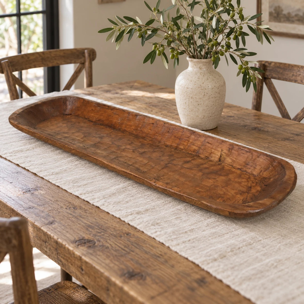 Extra-large wooden dough bowl styled empty on an oak dining table beside a cream vase and olive branches.