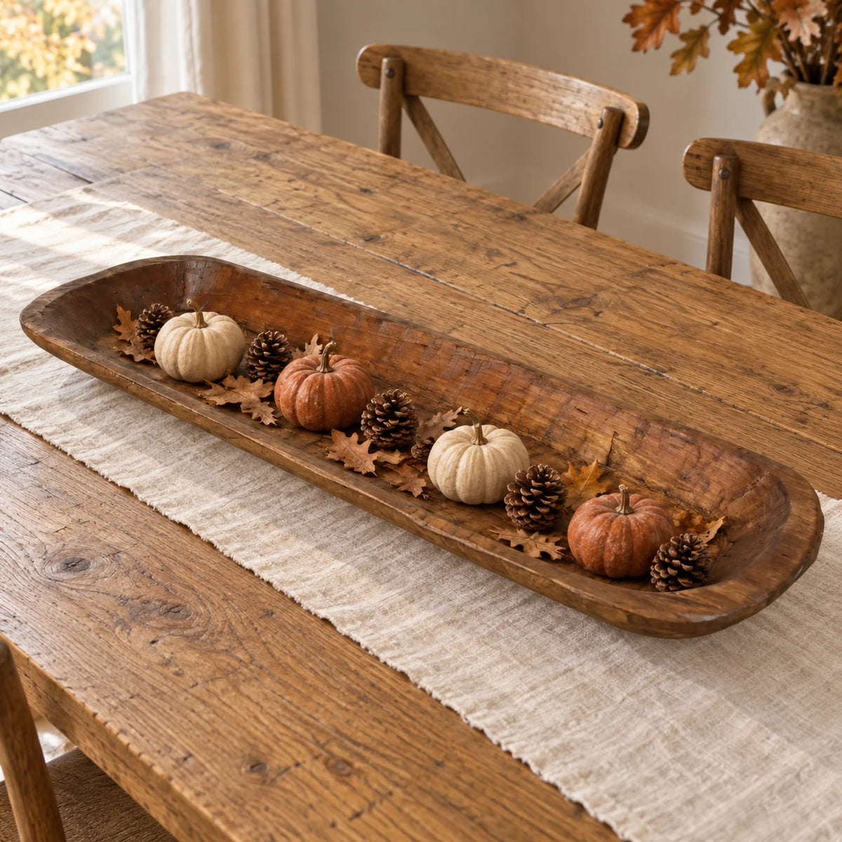 Extra-large wooden dough bowl styled with small pumpkins, pine cones and autumn leaves on a dining table.