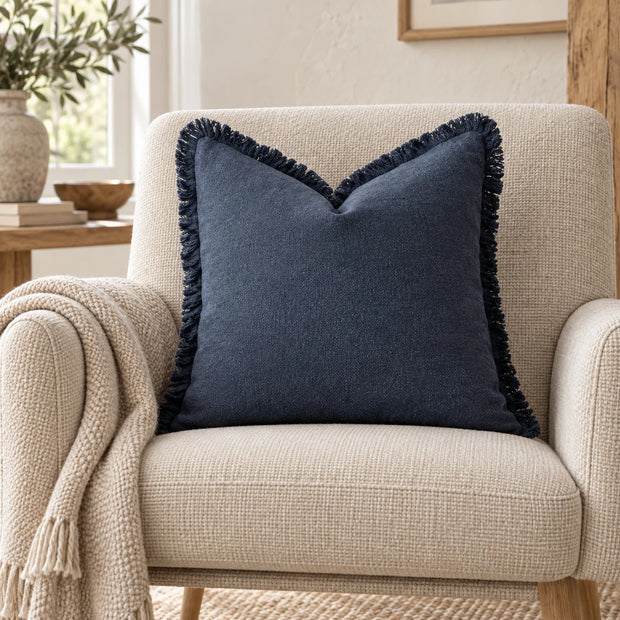 Navy blue frayed-edge cushion cover on an oatmeal armchair with a coordinating woven blanket.