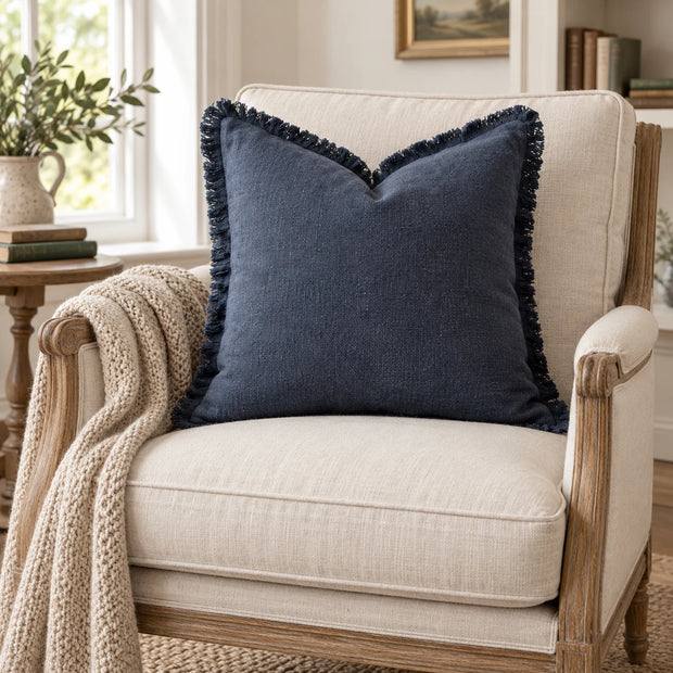 Navy blue frayed-edge cushion cover on an oak armchair with an oatmeal blanket in a reading corner.