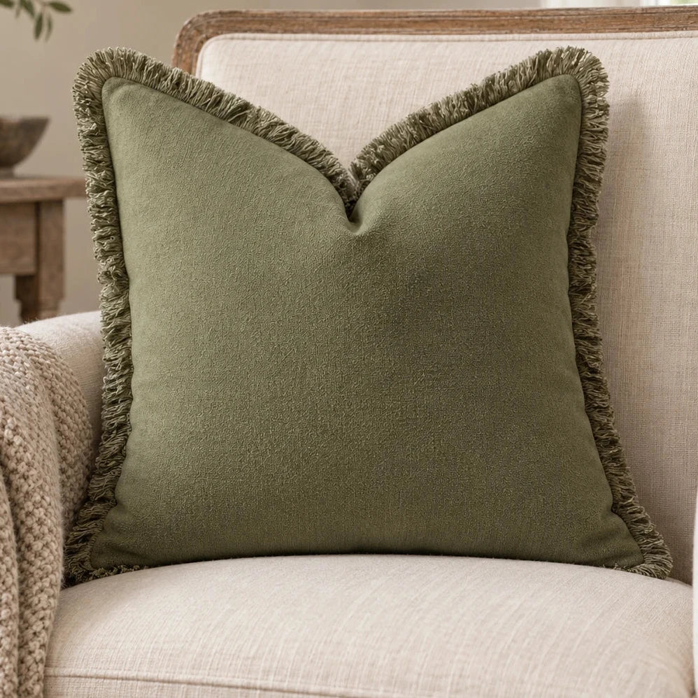 Frayed Edge Olive Green Cushion Cover on a cream wooden armchair beside an oatmeal knitted throw.