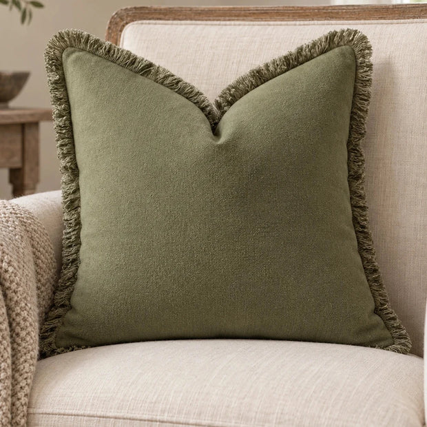 Frayed Edge Olive Green Cushion Cover on a cream wooden armchair beside an oatmeal knitted throw.
