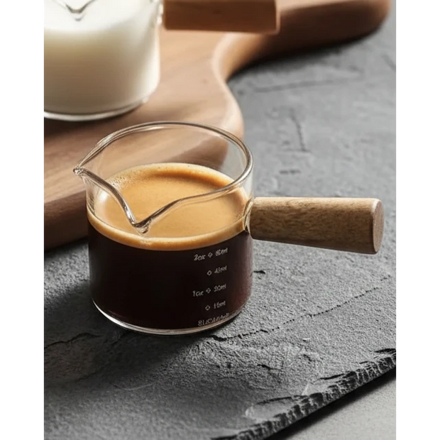 Glass Milk And Coffee Jug Pourer With Wooden Handle, clear glass jug filled with coffee, with a wooden handle on slate