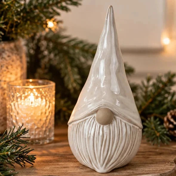 Small white glazed ceramic Christmas gonk on rustic wooden table by fir greenery and warm lights