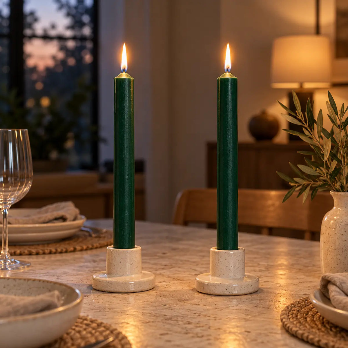Two green dinner candles burning in ceramic holders on a softly lit dining table.