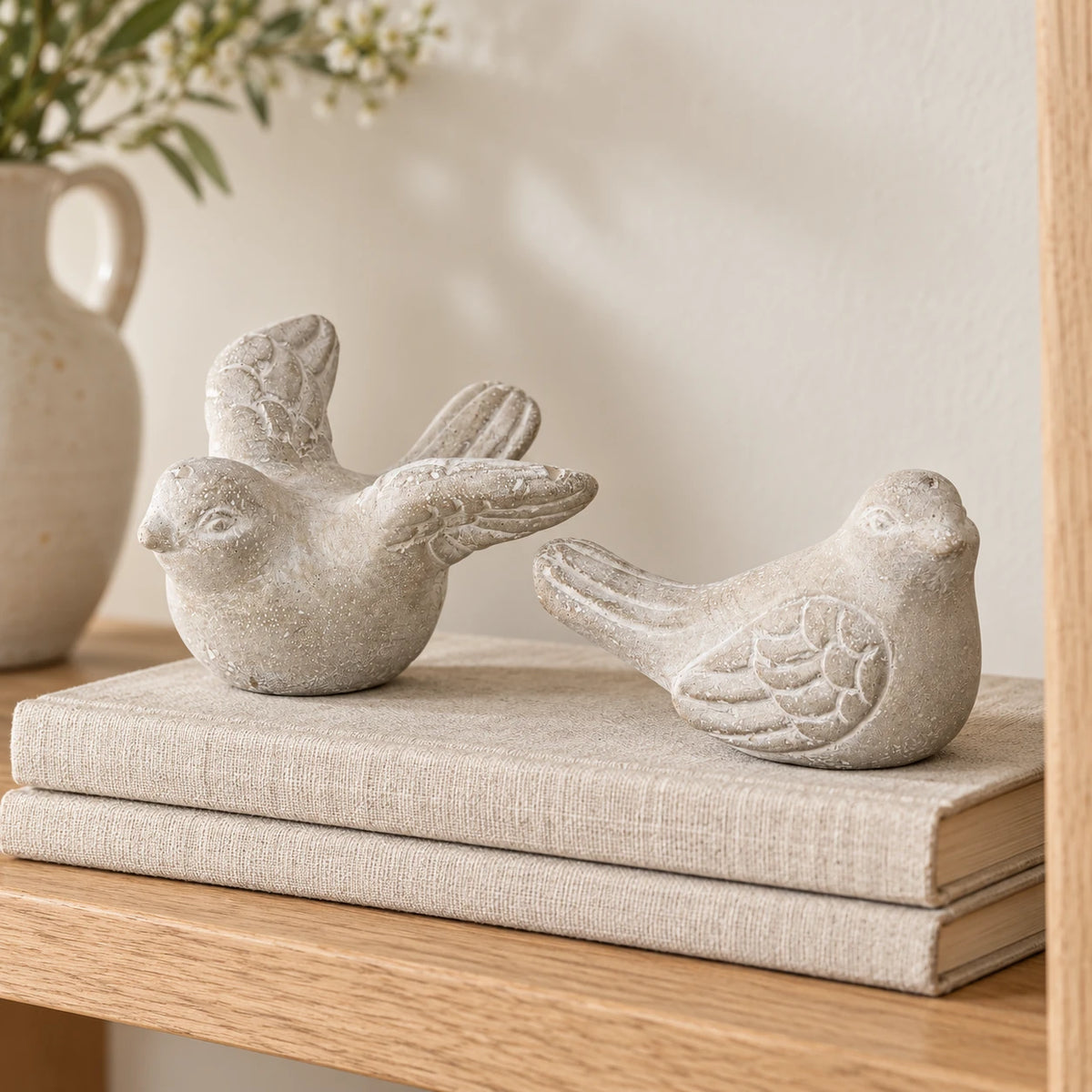 Pair of grey stone bird ornaments styled on oatmeal linen-covered books on an oak shelf.