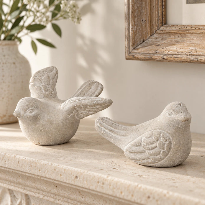 Grey stone birds, one with raised wings and one sitting, displayed together on a cream stone mantel.
