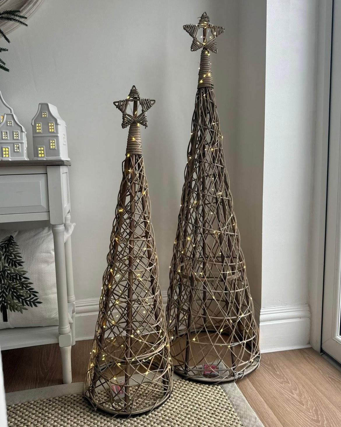 Two illuminated rattan Christmas trees with woven star toppers displayed beside a console; sold separately.