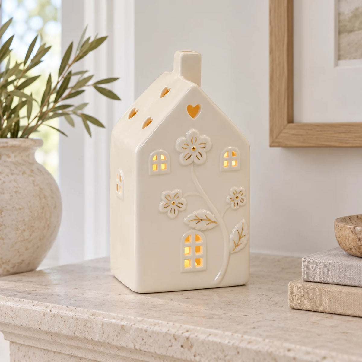 Large 17cm ivory ceramic LED house with raised flowers and gold-coloured details, mantel view, LED switched on.
