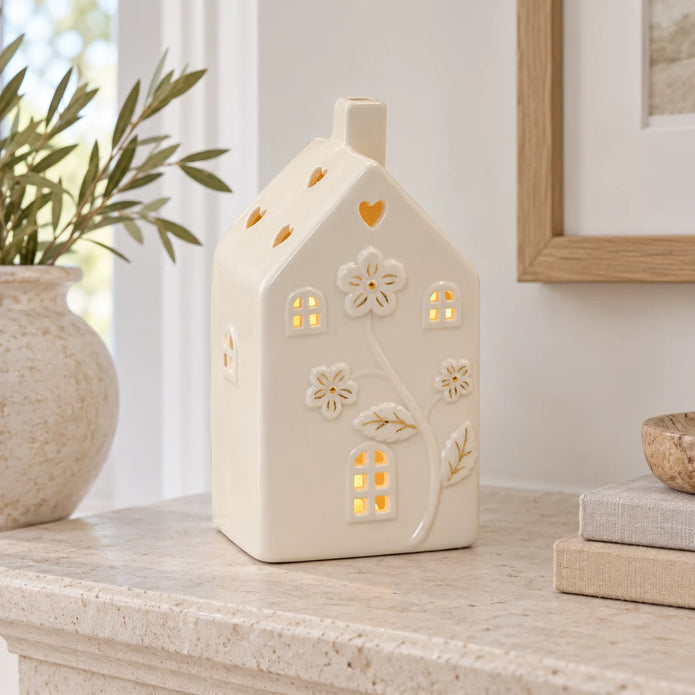 Large 17cm ivory ceramic LED house with raised flowers and gold-coloured details, mantel view, LED switched on.