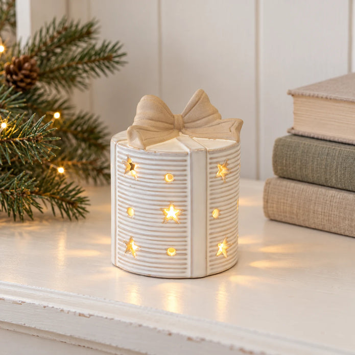 Ceramic light-up present with bow topper on a cream country-home shelf beside muted greenery.