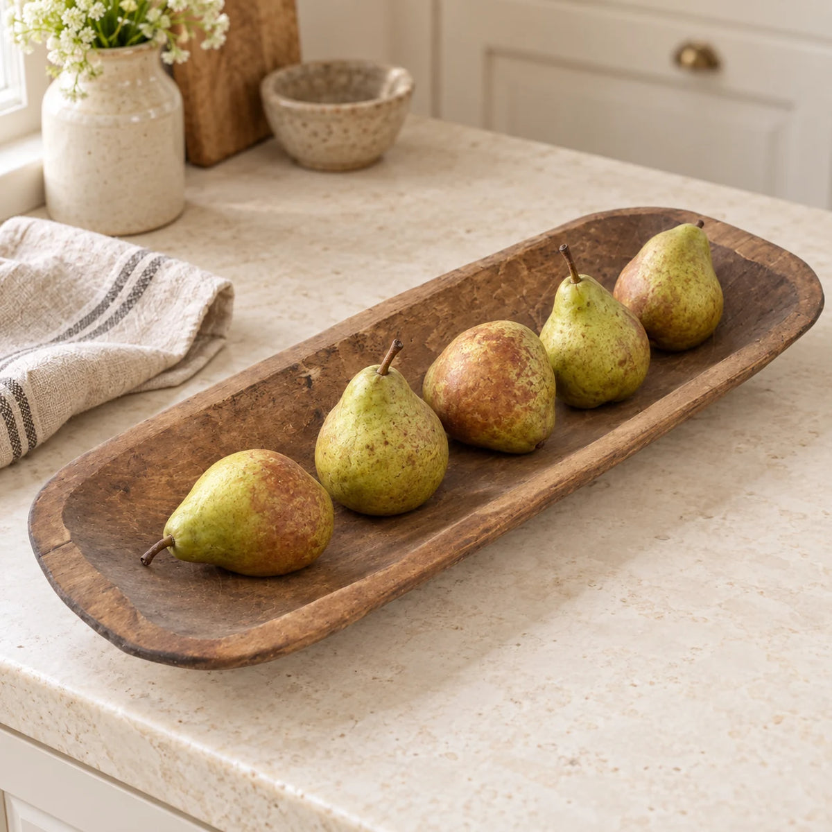 Large rustic wooden dough bowl holding green and russet pears on a cream kitchen worktop.
