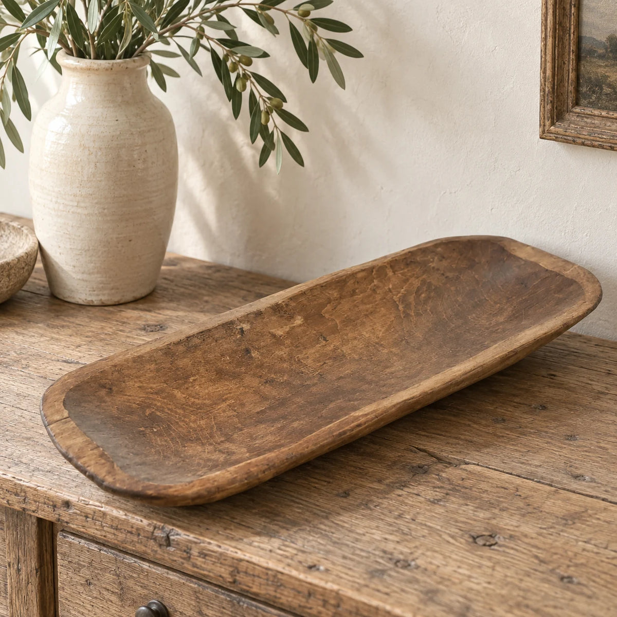 Large wooden dough bowl styled empty on an oak console beside a cream ceramic vase and foliage.