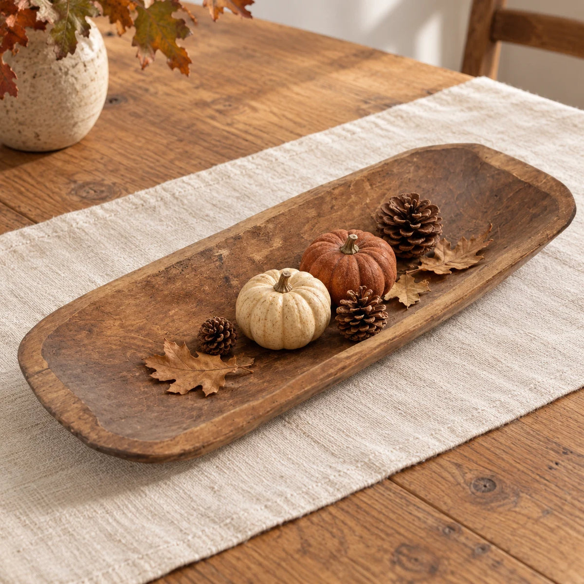 Large rustic dough bowl styled with small pumpkins, pine cones and autumn leaves on cream linen.