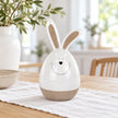 Large white ceramic rabbit with beige ears and base on a linen-covered oak dining table.