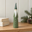 Slim green ceramic Christmas gonk beside cream books and winter foliage on an oak shelf.