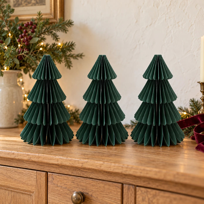 Three matching 25cm dark green paper Christmas trees on an oak sideboard with festive greenery.