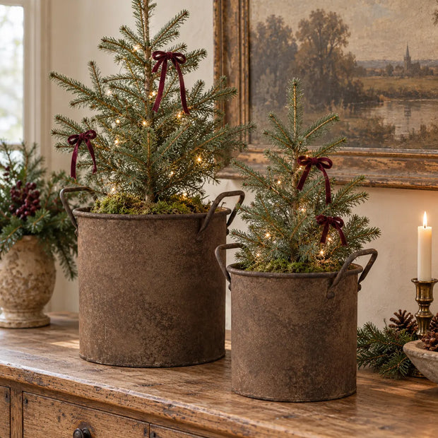 Large and small rust metal planter pots holding miniature Christmas trees on an antique oak console