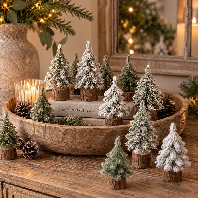 Miniature green and snowy Christmas trees on rustic log bases styled in a wooden bowl with festive candles