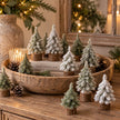 Miniature green and snowy Christmas trees on rustic log bases styled in a wooden bowl with festive candles