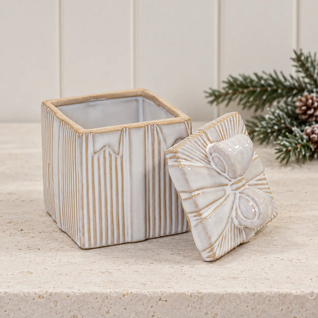 Open natural ceramic gift box with removable bow lid beside evergreen foliage
