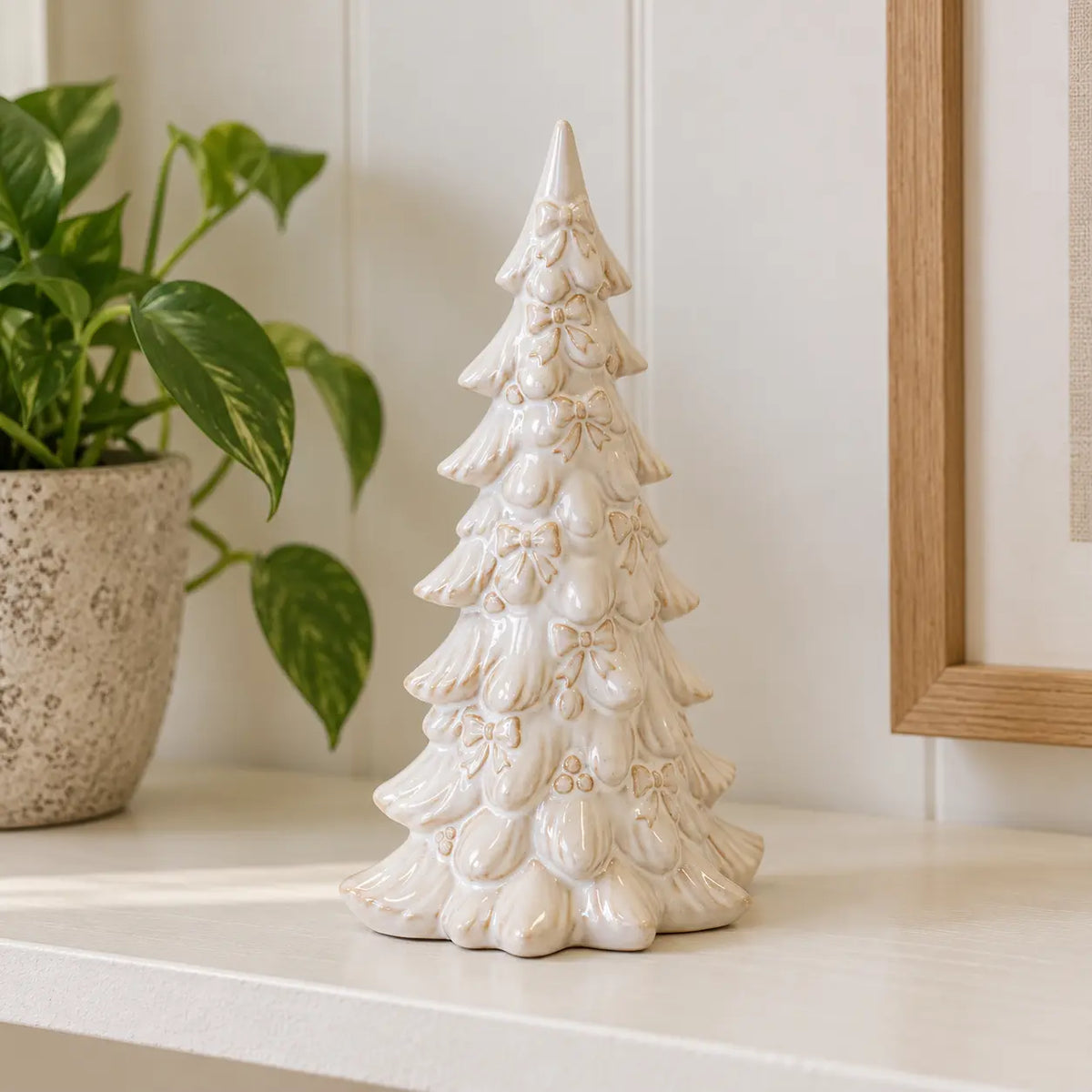 Small ivory layered ceramic Christmas tree on a neutral shelf beside a houseplant.