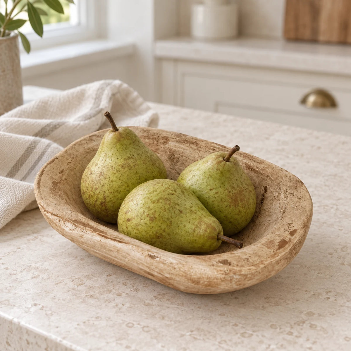 Natural pine mini dough bowl holding three green pears on a cream kitchen counter beside linen.
