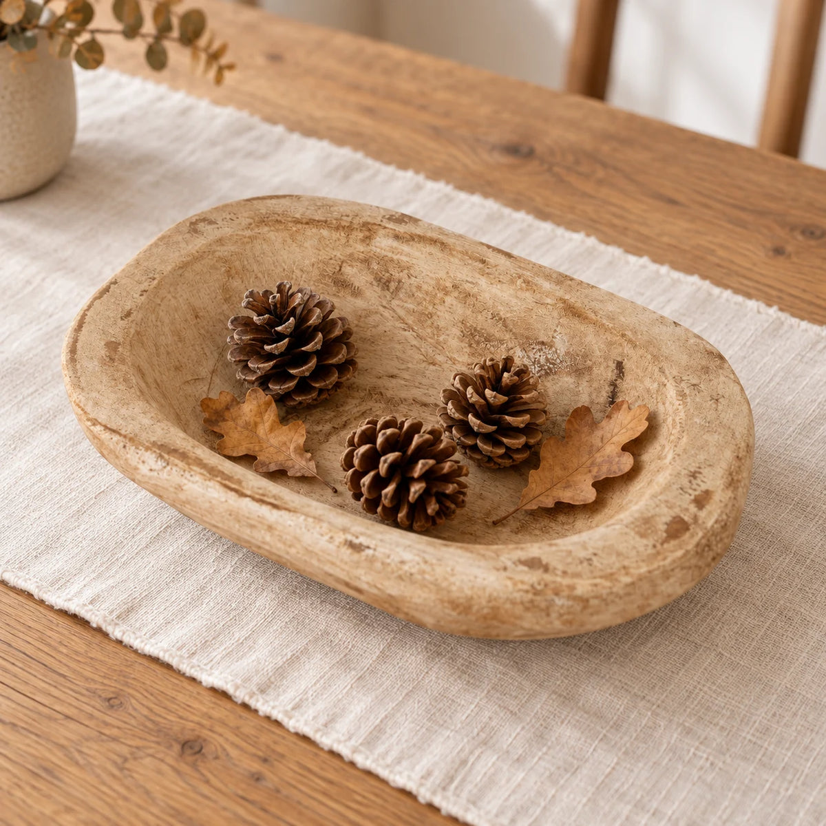 Natural pine mini dough bowl with pine cones and dried oak leaves on a cream linen runner.