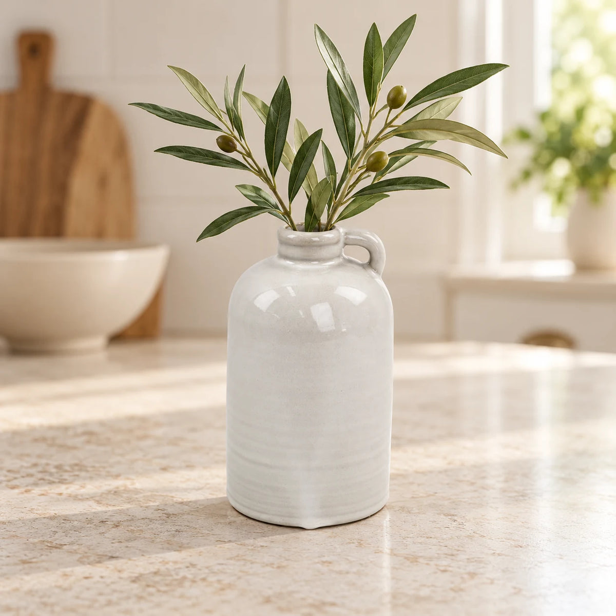 Short porcelain jug vase with olive foliage on a bright cream kitchen countertop
