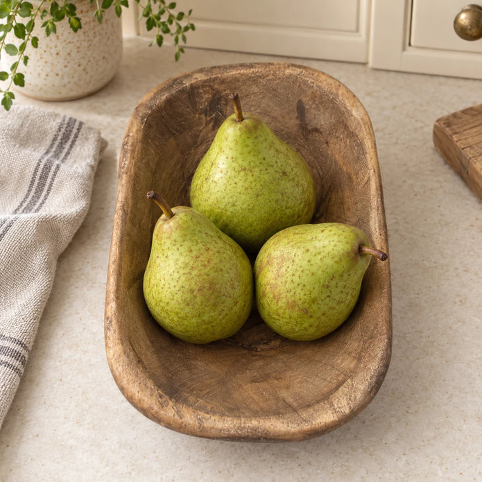 Natural walnut mini dough bowl holding three pears on a cream kitchen worktop beside oatmeal linen.