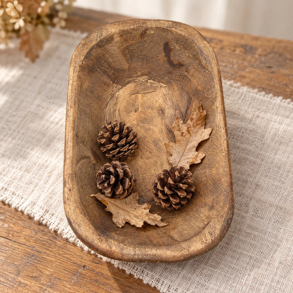Natural walnut mini dough bowl containing pine cones and dried leaves on a cream linen runner.