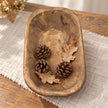 Natural walnut mini dough bowl containing pine cones and dried leaves on a cream linen runner.