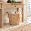 Natural woven basket with handles storing cream scarves beneath a wooden hallway console