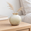 Medium round neutral ceramic bud vase with delicate dried grasses on an oak bedside table.