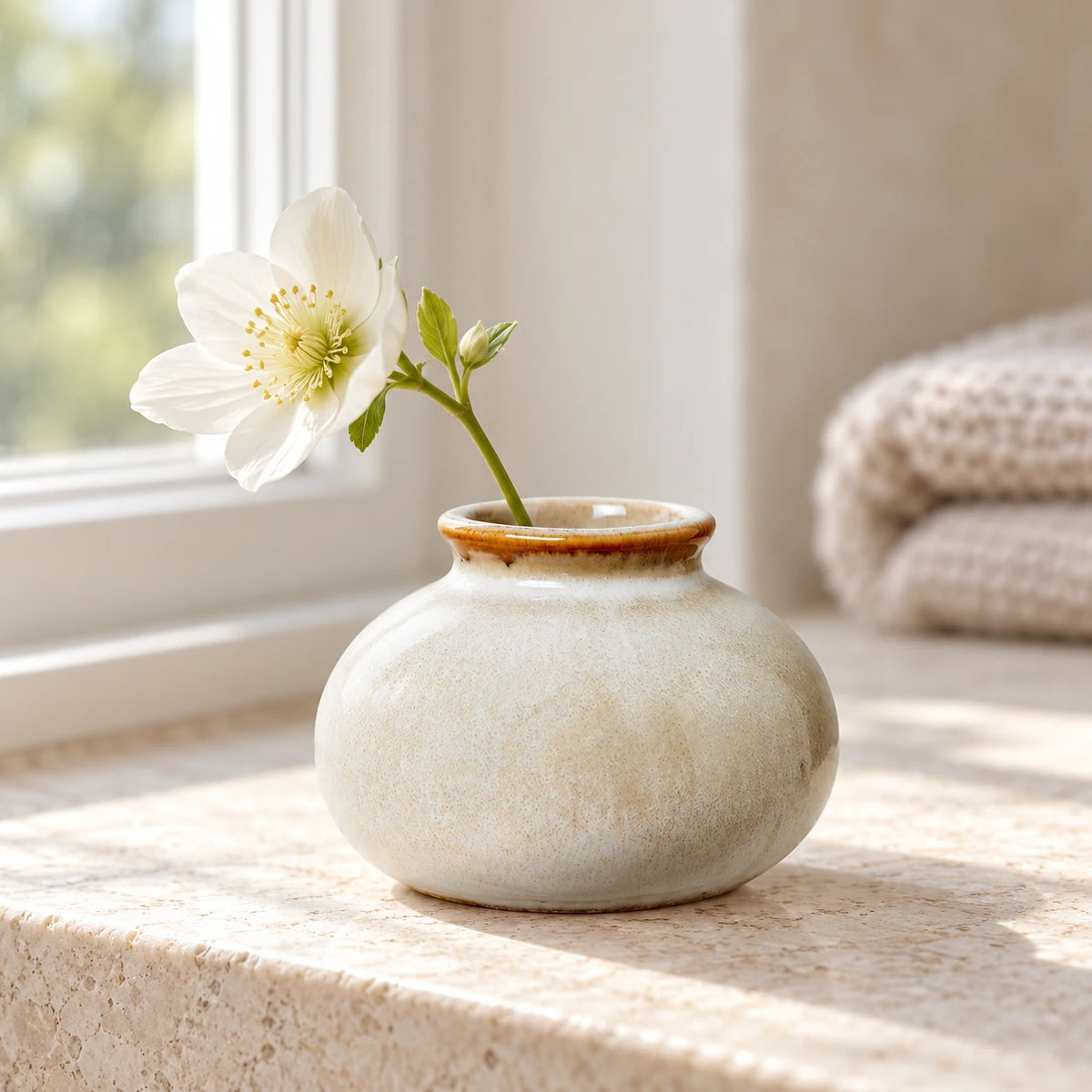 Small cream ceramic bud vase with a white flower on a light stone windowsill.