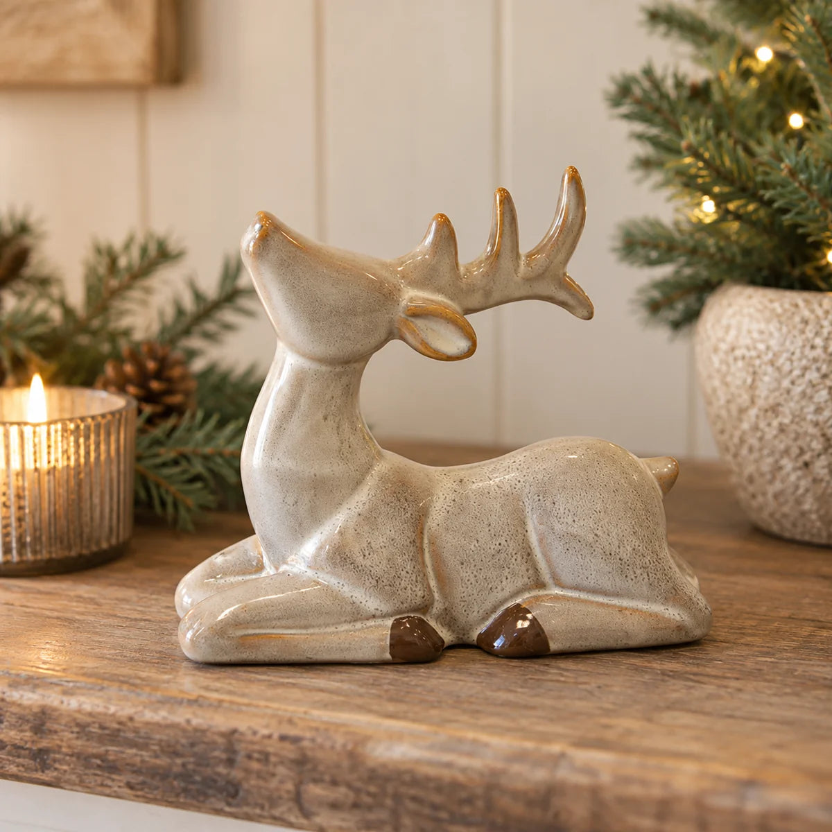 Reclining neutral ceramic reindeer with raised antlers displayed beside evergreen branches and candle