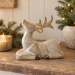 Reclining neutral ceramic reindeer with raised antlers displayed beside evergreen branches and candle