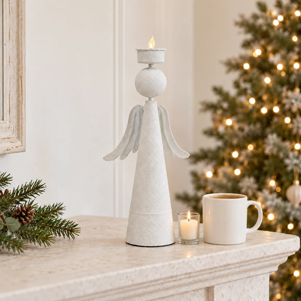 32cm off-white metal angel tealight holder on a Christmas mantel beside a coffee-filled mug for scale and a small candle in a glass holder.