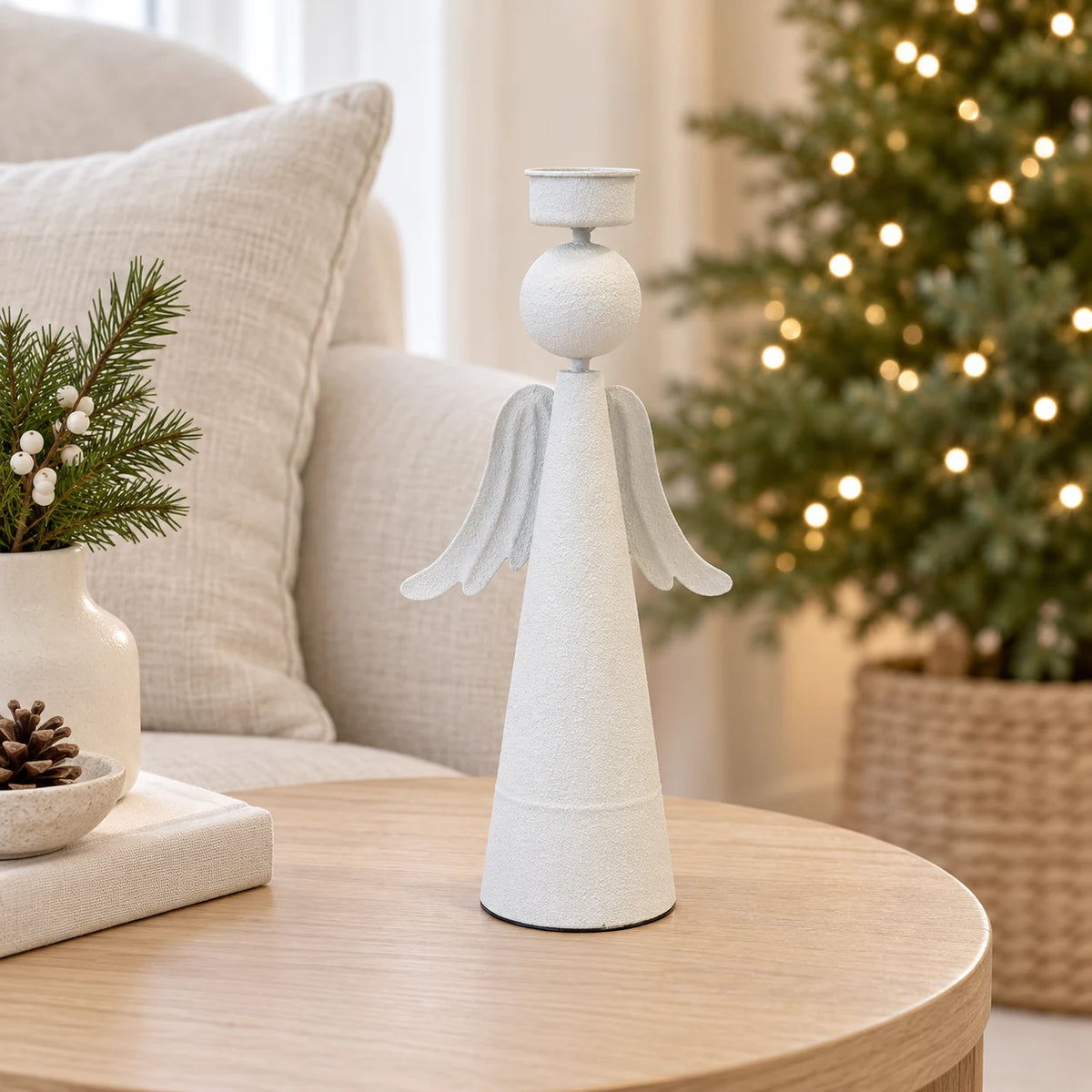 Empty off-white metal angel tealight holder on an oak side table with a softly lit Christmas tree in the background.