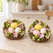 Set of two decorative pastel egg nests with pink flowers and greenery, packaging removed, table view.