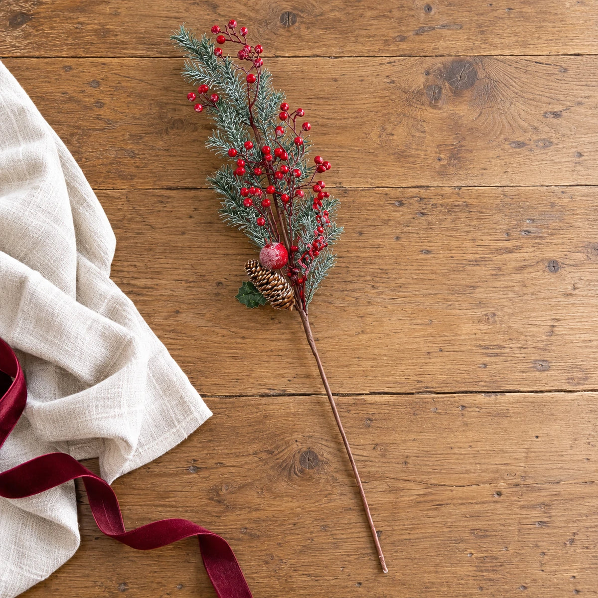 Full red berry and cedar festive stem laid on an oak table beside cream linen.