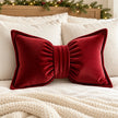 Red bow-shaped corduroy cushion styled on ivory bedding beneath a Christmas foliage garland.