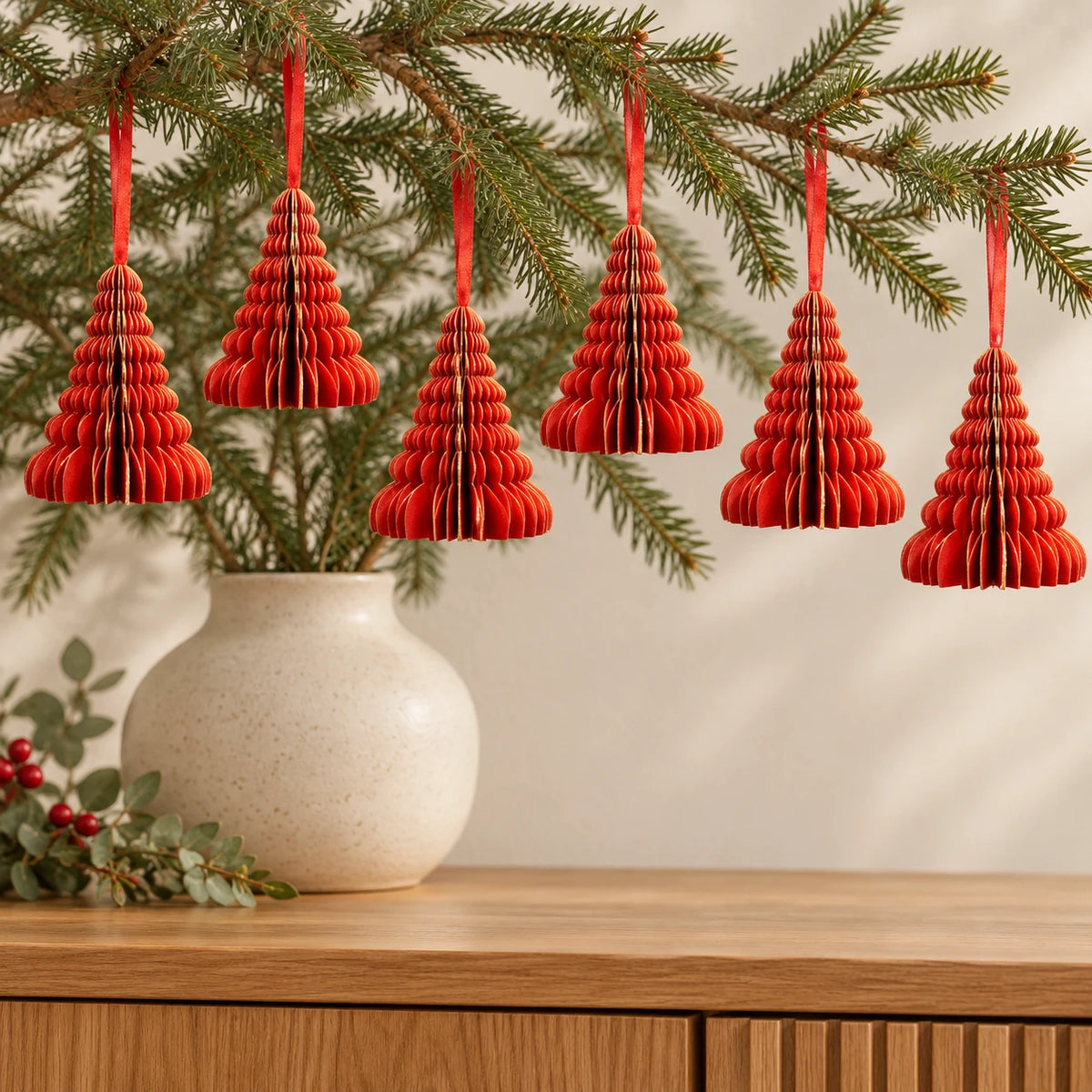 Six red paper tree decorations with gold edges hanging from fir branches above an oak sideboard.