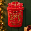 Red metal North Pole Express Mail letterbox against a dark green wall with festive greenery and a wrapped gift.